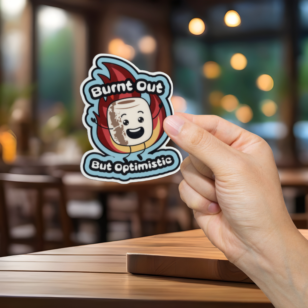 Hand holding a sticker with a cartoon marshmallow character and text 'Burnt Out But Optimistic' in a blurred indoor setting.