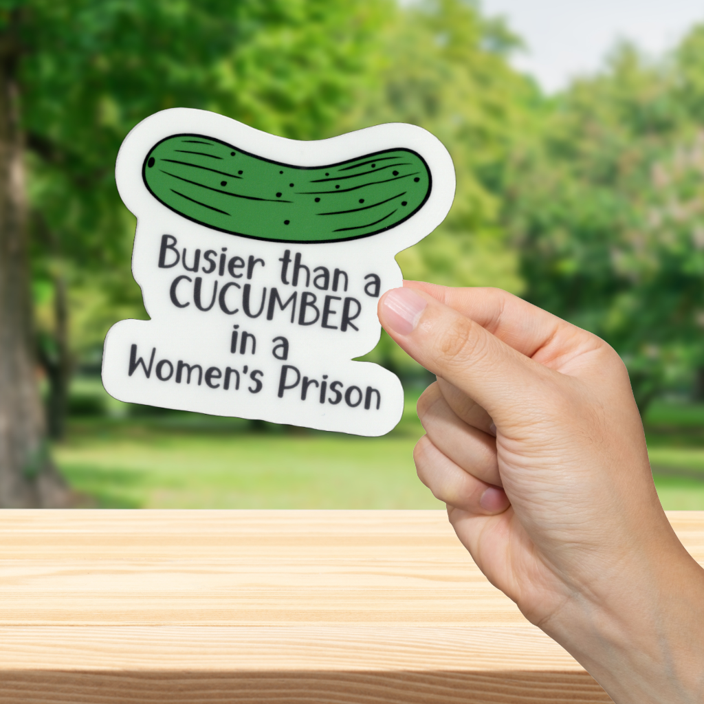 Hand holding a sticker with a pickle graphic and text in front of a blurred green outdoor background