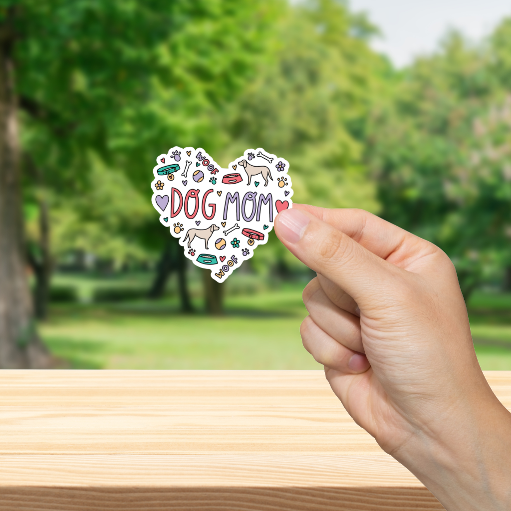 Heart-shaped 'DOG MOM' sticker held by a hand with a blurred green outdoor background