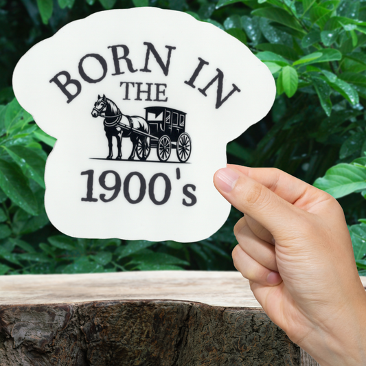 Hand holding a sign with 'Born in the 1900's' and a horse-drawn carriage design against a green leafy background.
