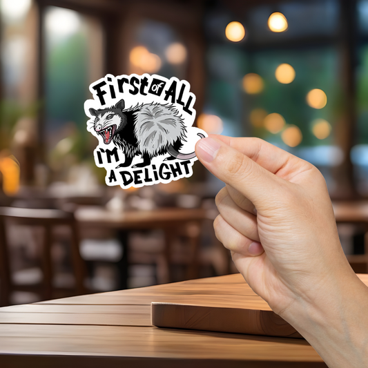 Hand holding a sticker with a cartoon opossum and text 'First of all, I'm a delight' in a blurred restaurant setting