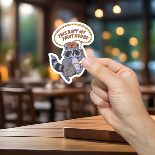 Hand holding a sticker with a raccoon graphic and text 'This ain't my first rodeo' in a blurred restaurant setting