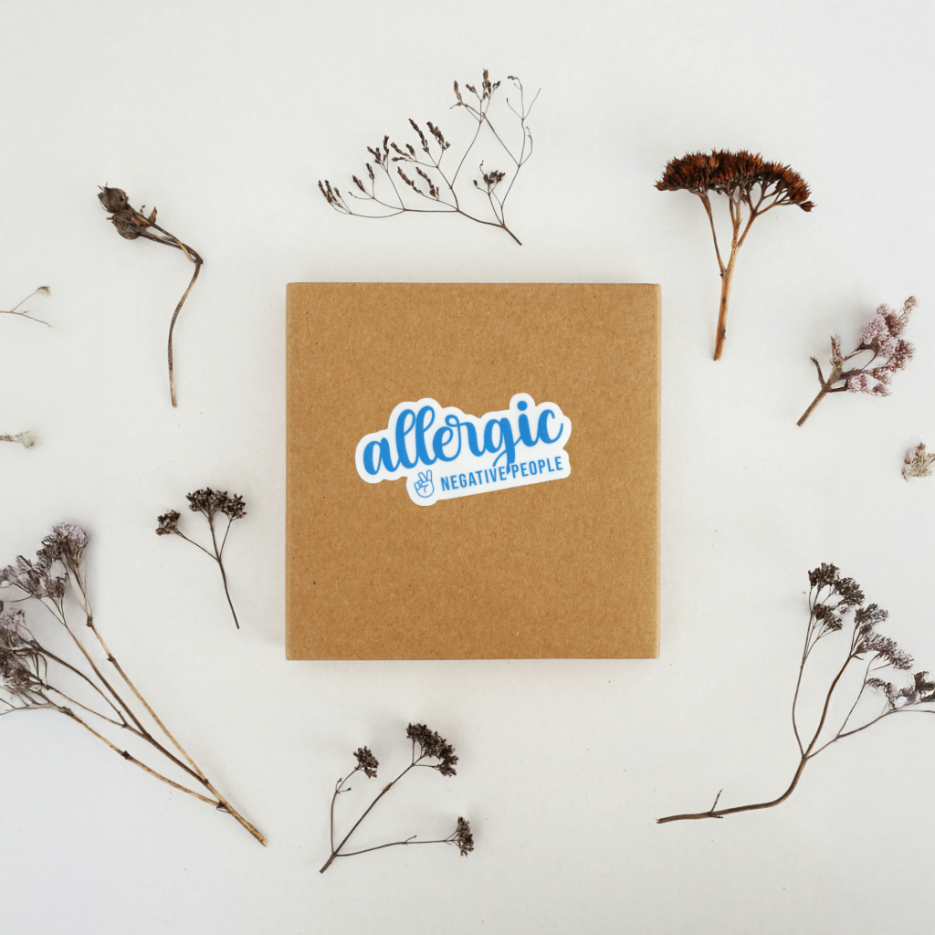 Brown square card with 'allergic to negative people' sticker surrounded by dried flowers on a light background