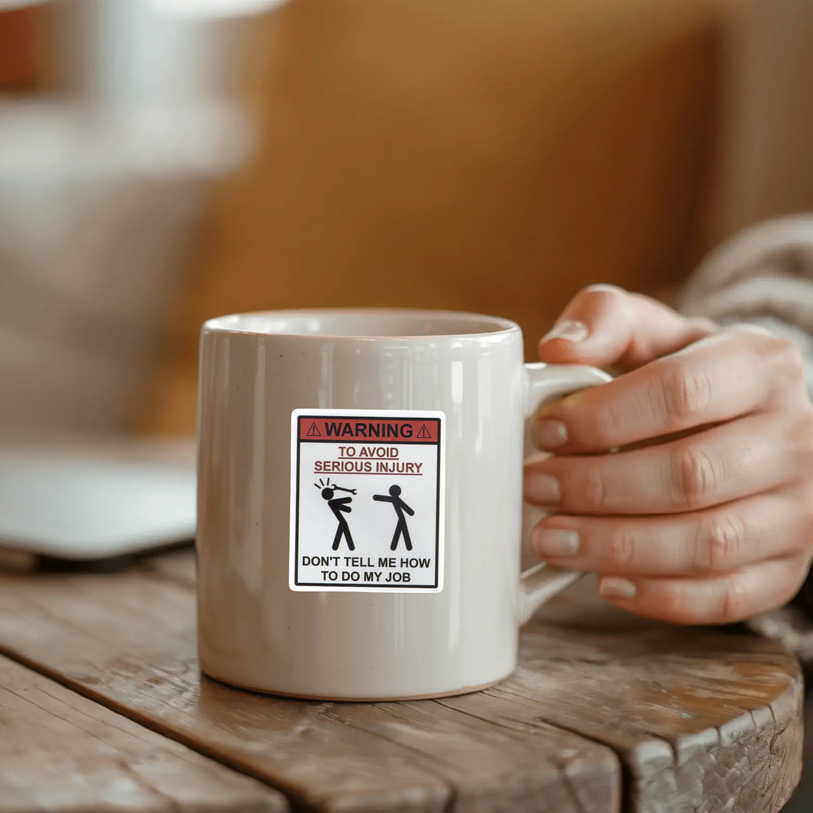 Person holding a mug with a humorous Avoid Injury Sticker warning label on a wooden surface