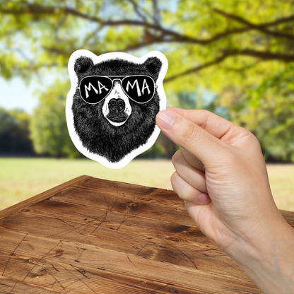 Hand holding a sticker of a bear wearing sunglasses with 'MAMA' text against a blurred outdoor background.