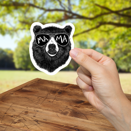 Hand holding a sticker of a bear wearing sunglasses with 'MAMA' text against a blurred outdoor background.