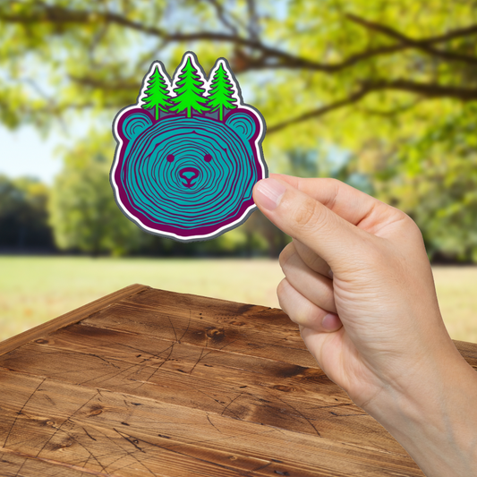 Hand holding a sticker with a bear design and trees against a blurred outdoor background