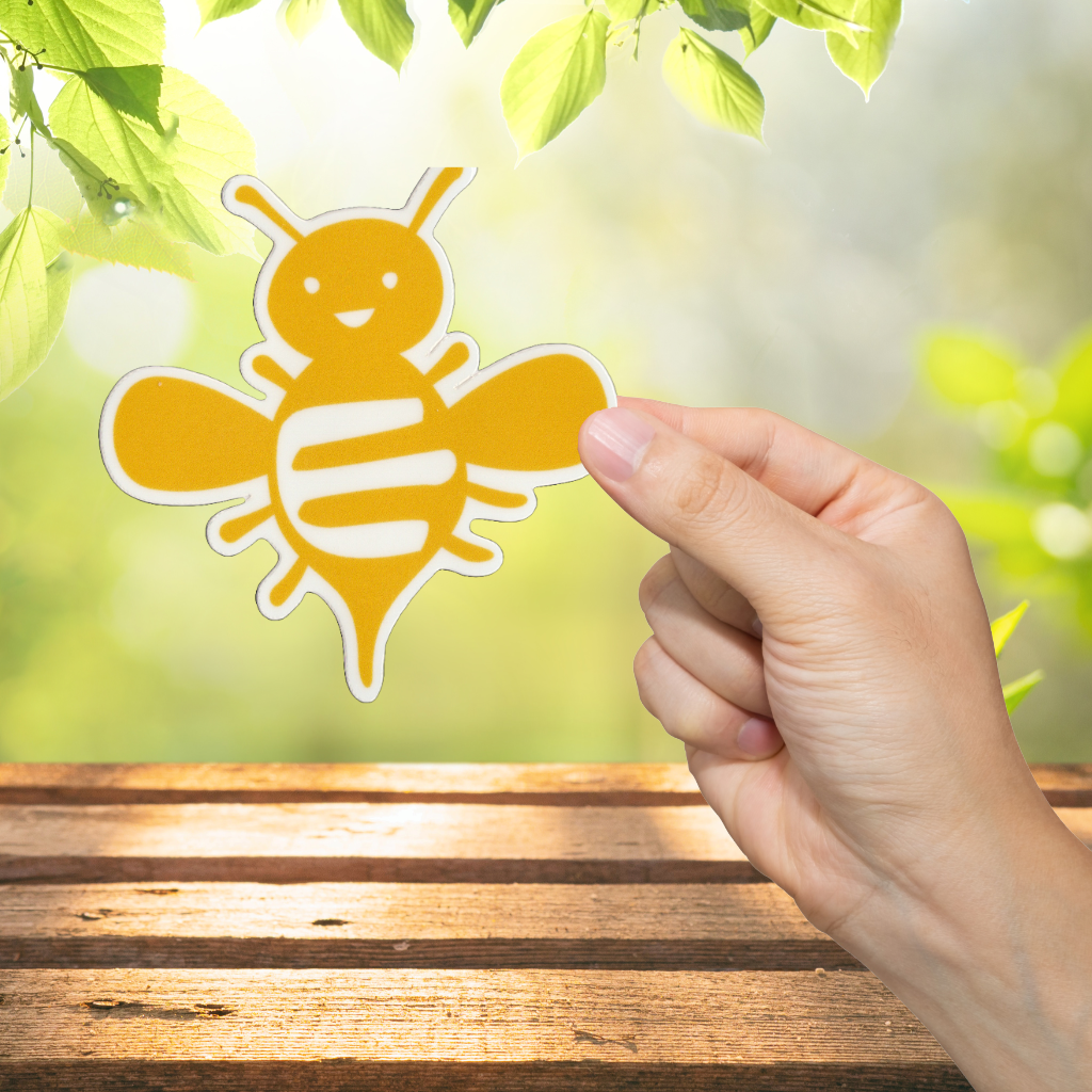 Hand holding a yellow bee-shaped sticker against a natural background with green leaves.