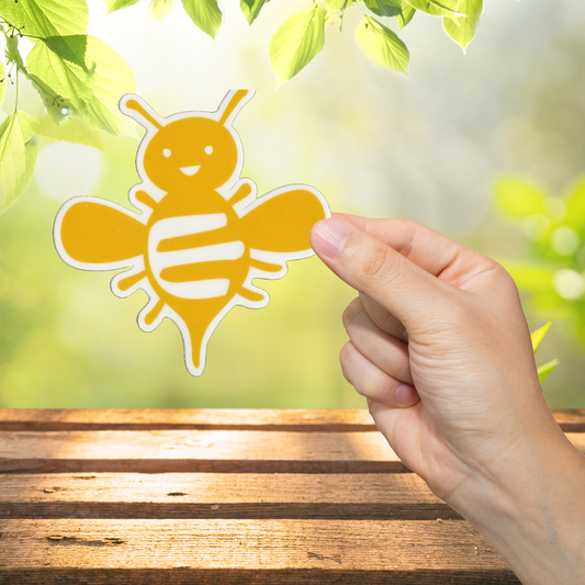 Hand holding a yellow bee-shaped sticker against a natural background with green leaves.