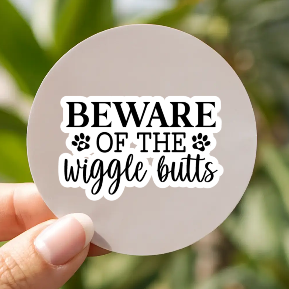 Circular sign with 'Beware of the wiggle butts' text held by a hand against a blurred green outdoor background