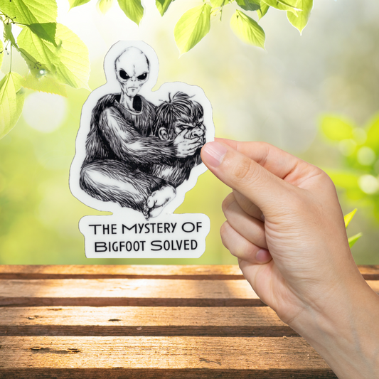 Sticker with alien holding a sasquatch mask with text 'The Mystery of Bigfoot Solved', held by a hand on a wooden surface with greenery in the background.