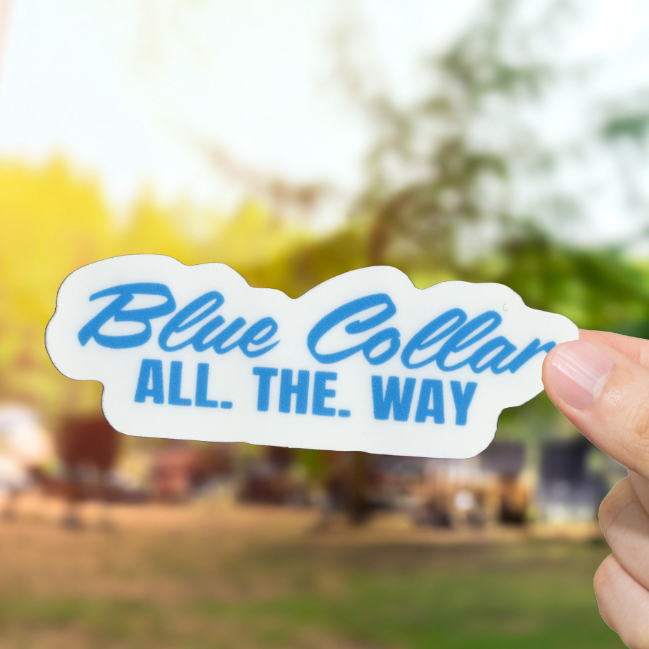 Hand holding a 'Blue Collar All. The. Way' sticker with a blurred outdoor background