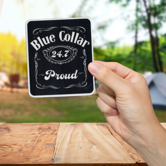 Hand holding a sign with 'Blue Collar 24.7 Proud' text against a blurred outdoor background.