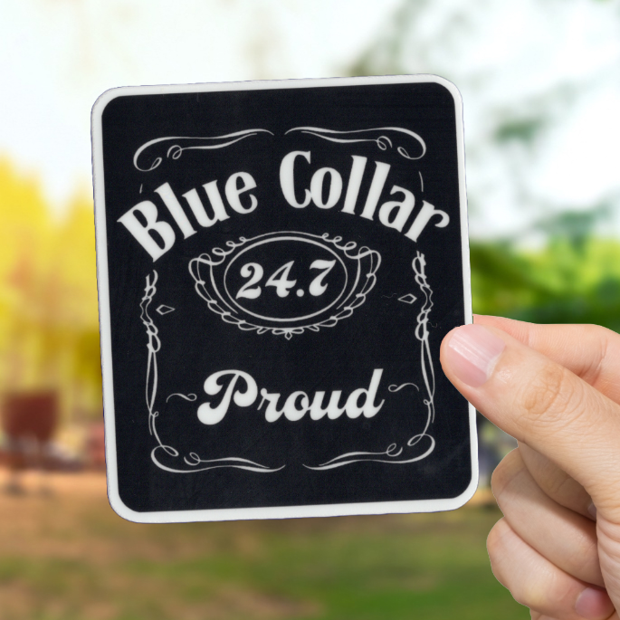Hand holding a sign with 'Blue Collar 24.7 Proud' text against a blurred outdoor background.
