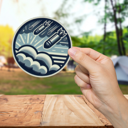 Hand holding a circular sticker with a design of missiles and waves on a wooden surface with a blurred natural background.