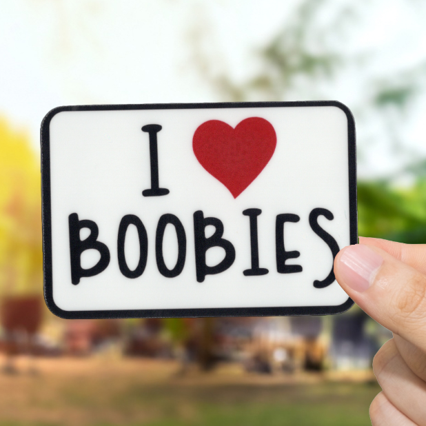 Hand holding a sign with 'I ❤️ BOOBIES' text outdoors.