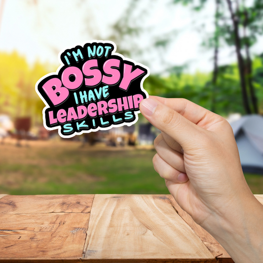 Hand holding a colorful sticker with text 'I'm not bossy, I have leadership skills' against a blurred outdoor background.