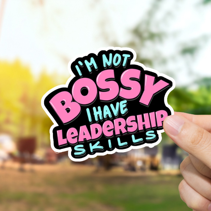Hand holding a colorful sticker with text 'I'm not bossy, I have leadership skills' against a blurred outdoor background.