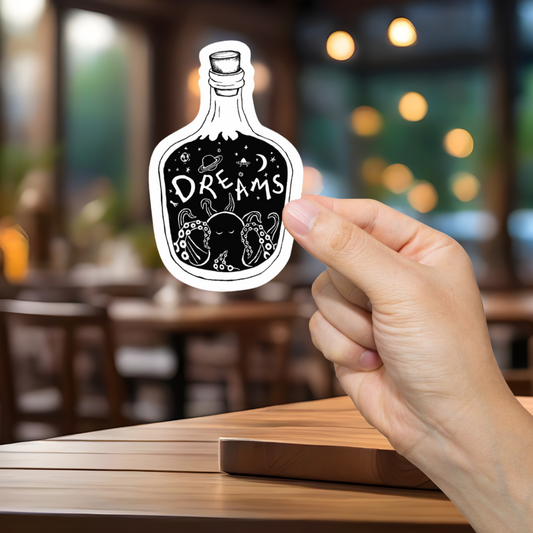 Hand holding a sticker of a bottle labeled 'Dreams' in a blurred restaurant setting