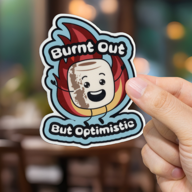 Hand holding a sticker with a cartoon marshmallow character and text 'Burnt Out But Optimistic' in a blurred indoor setting.