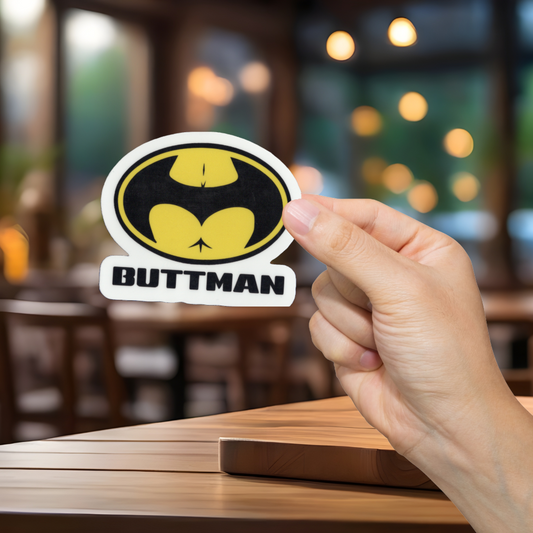 Hand holding a sticker with a bat symbol and 'BUTTMAN' text in a blurred indoor setting.