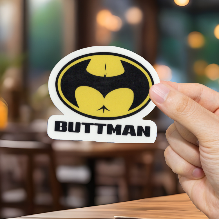 Hand holding a sticker with a bat symbol and 'BUTTMAN' text in a blurred indoor setting.