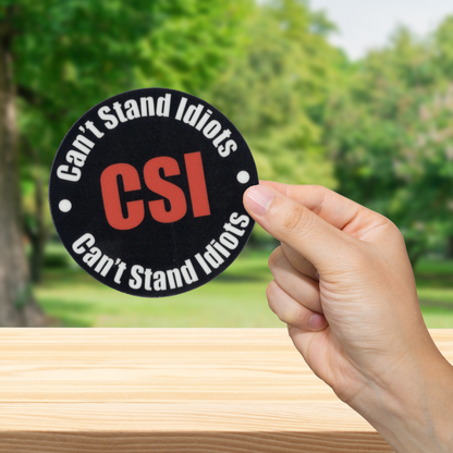 Hand holding a 'Can't Stand Idiots CSI' sticker with a blurred green outdoor background