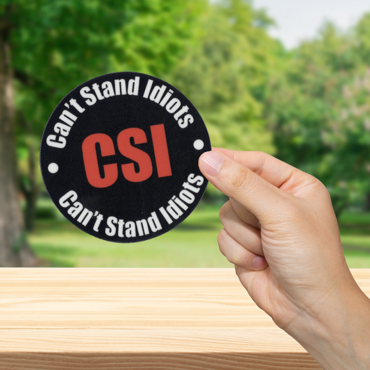 Hand holding a 'Can't Stand Idiots CSI' sticker with a blurred green outdoor background