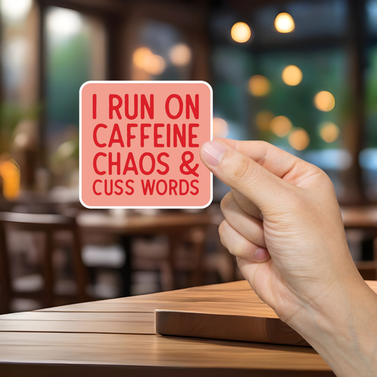 Hand holding a sign with text 'I Run on Caffeine, Chaos & Cuss Words' in a blurred cafe setting
