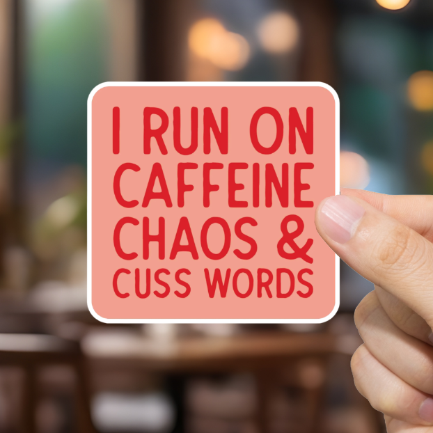 Hand holding a sign with text 'I Run on Caffeine, Chaos &amp; Cuss Words' in a blurred cafe setting
