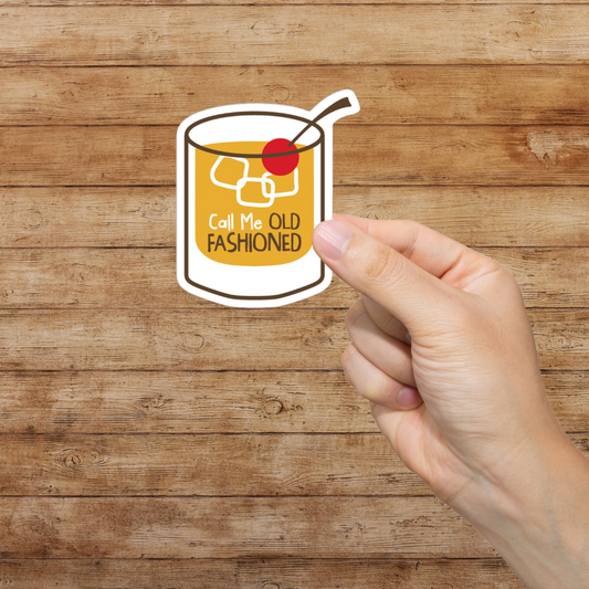 Hand holding a sticker of an old fashioned cocktail with text on a wooden background. Call me old fashioned.