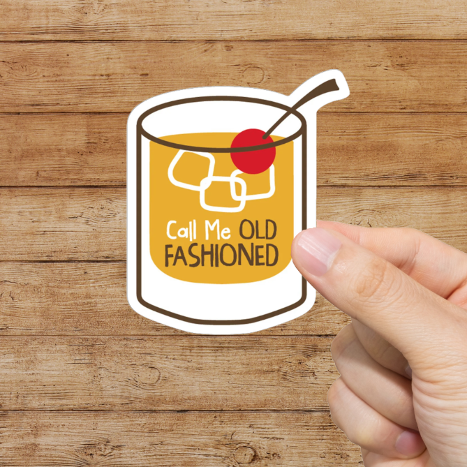 Hand holding a sticker of an old fashioned cocktail with text on a wooden background. Call me old fashioned.