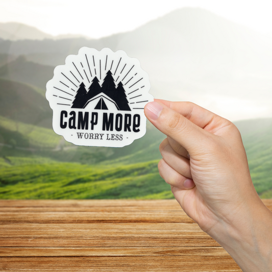 Hand holding a 'Camp More Worry Less' sticker with a scenic background