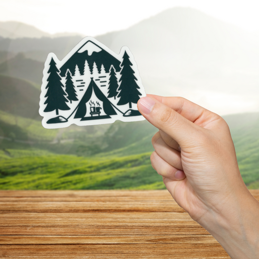 Hand holding a sticker of a tent and trees against a scenic background