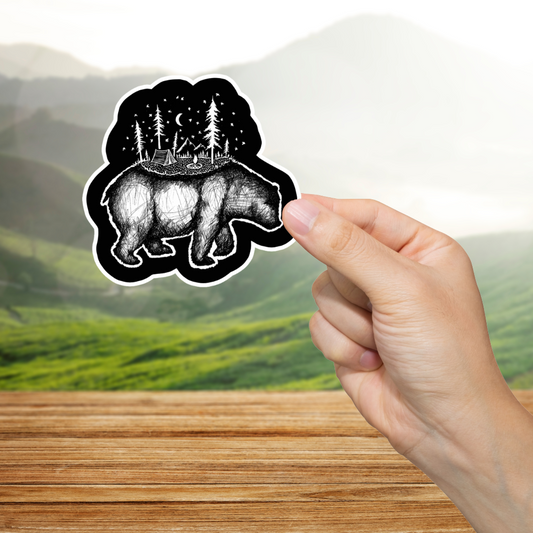 Hand holding a black and white bear sticker with a forest design against a blurred natural background