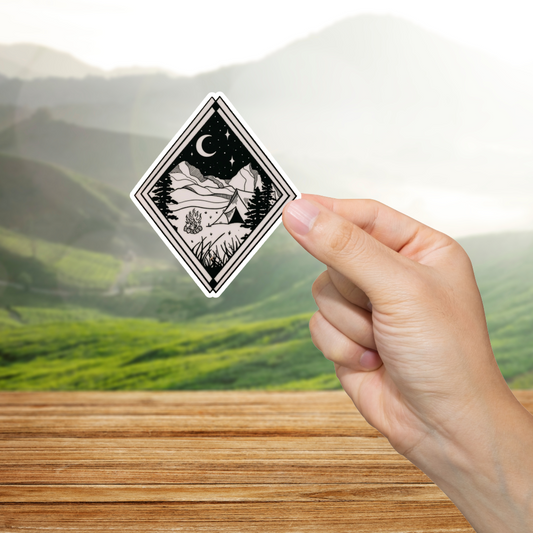 Hand holding a black and white diamond-shaped sticker with a nature design against a blurred mountain background.