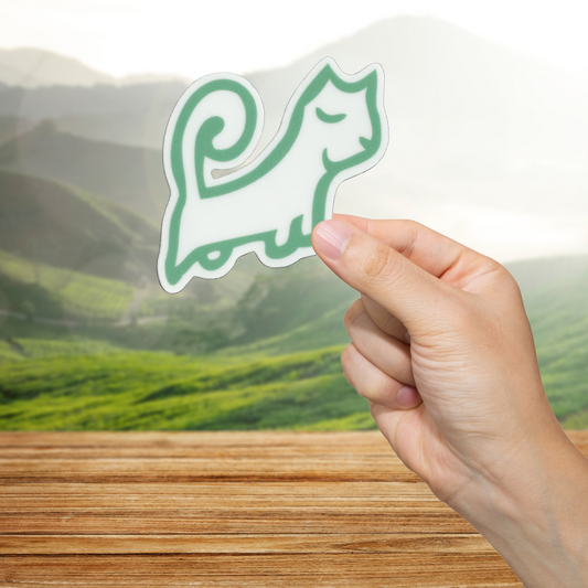 Hand holding a green cat-shaped sticker with a scenic background