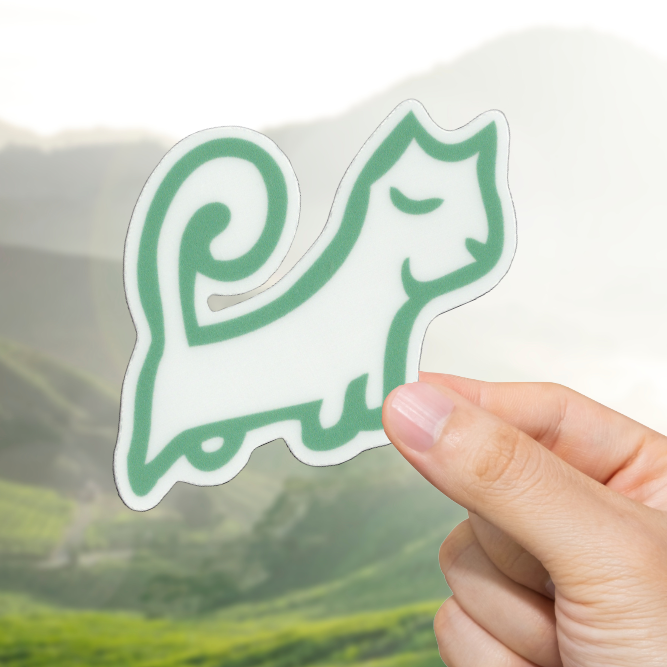Hand holding a green cat-shaped sticker with a scenic background