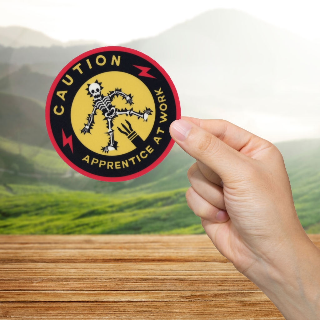 Hand holding a 'Caution: Apprentice at Work' sticker with a scenic background