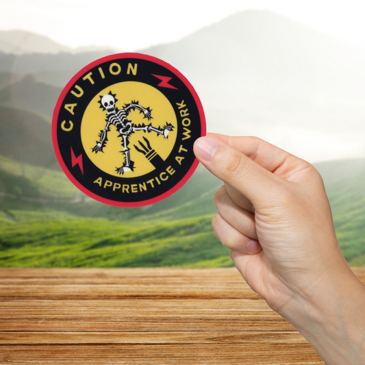Hand holding a 'Caution: Apprentice at Work' sticker with a scenic background