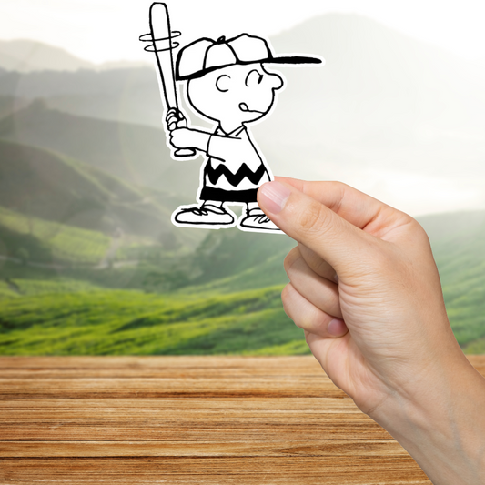 Hand holding a sticker of Charlie Brown with a baseball bat against a scenic background