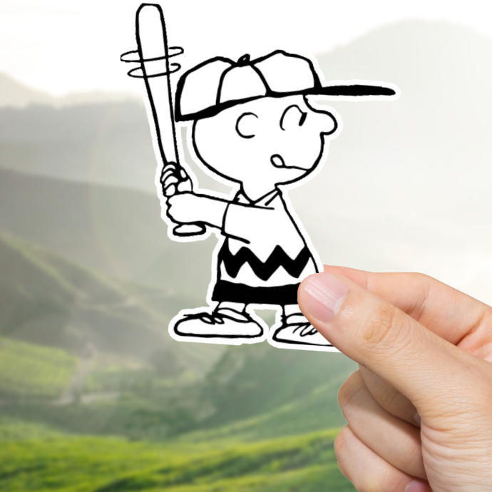 Hand holding a sticker of Charlie Brown with a baseball bat against a scenic background