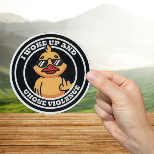 Sticker with a duck wearing sunglasses and text 'I woke up and chose violence' held by a hand on a wooden surface with a scenic background.