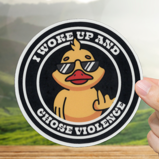 Sticker with a duck wearing sunglasses and text 'I woke up and chose violence' held by a hand on a wooden surface with a scenic background.