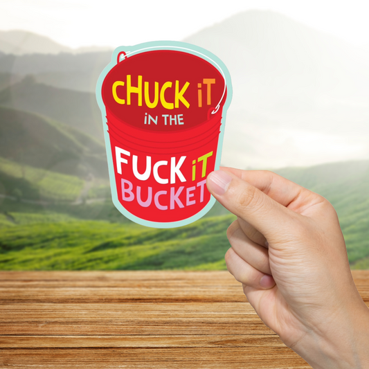 Hand holding a red bucket-shaped sticker with text "Fuck It Bucket" against a scenic background