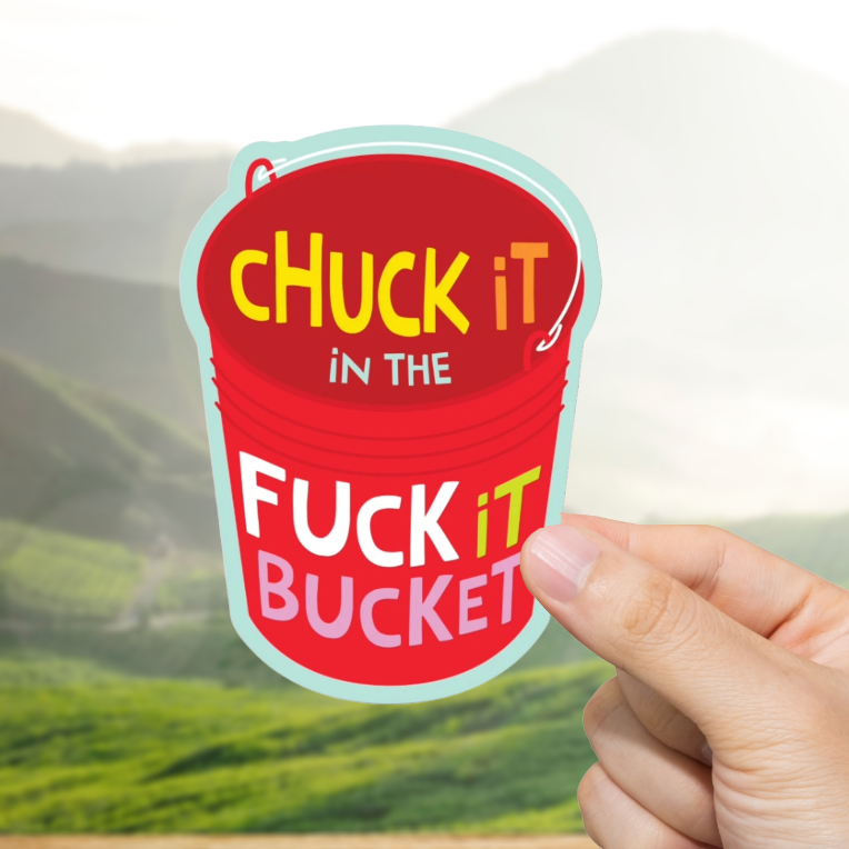Hand holding a red bucket-shaped sticker with text "Fuck It Bucket" against a scenic background