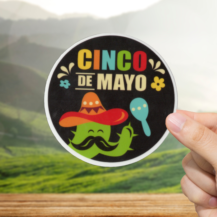 Hand holding a 'Cinco de Mayo' sticker with a colorful design on a scenic background.
