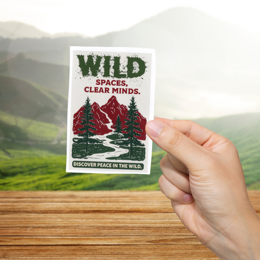 Hand holding a sticker with 'Wild Spaces, Clear Minds' text and mountain design against a scenic background.
