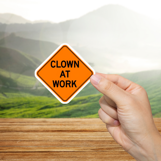 Hand holding a sign that reads 'Clown at Work' with a scenic background