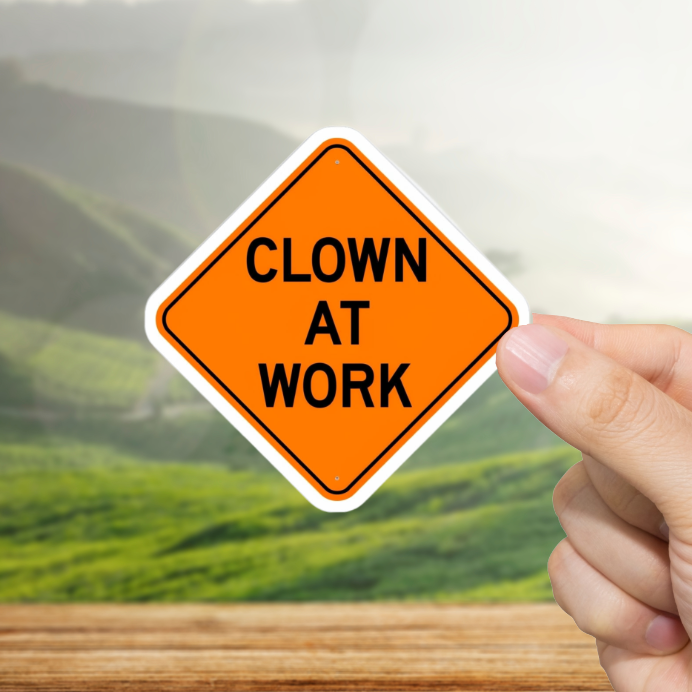 Hand holding a sign that reads 'Clown at Work' with a scenic background
